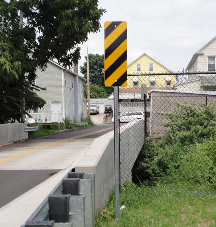 Black and yellow diagonal striped warning sign on a post beside a narrow bridge installed by Minichi Inc.