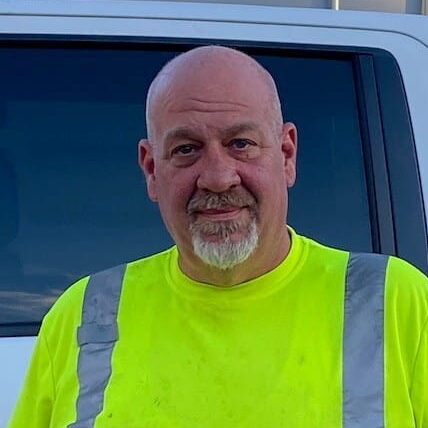 Man named Chris Nelson with a shaved head and goatee wearing a high-visibility yellow shirt stands in front of a white vehicle.