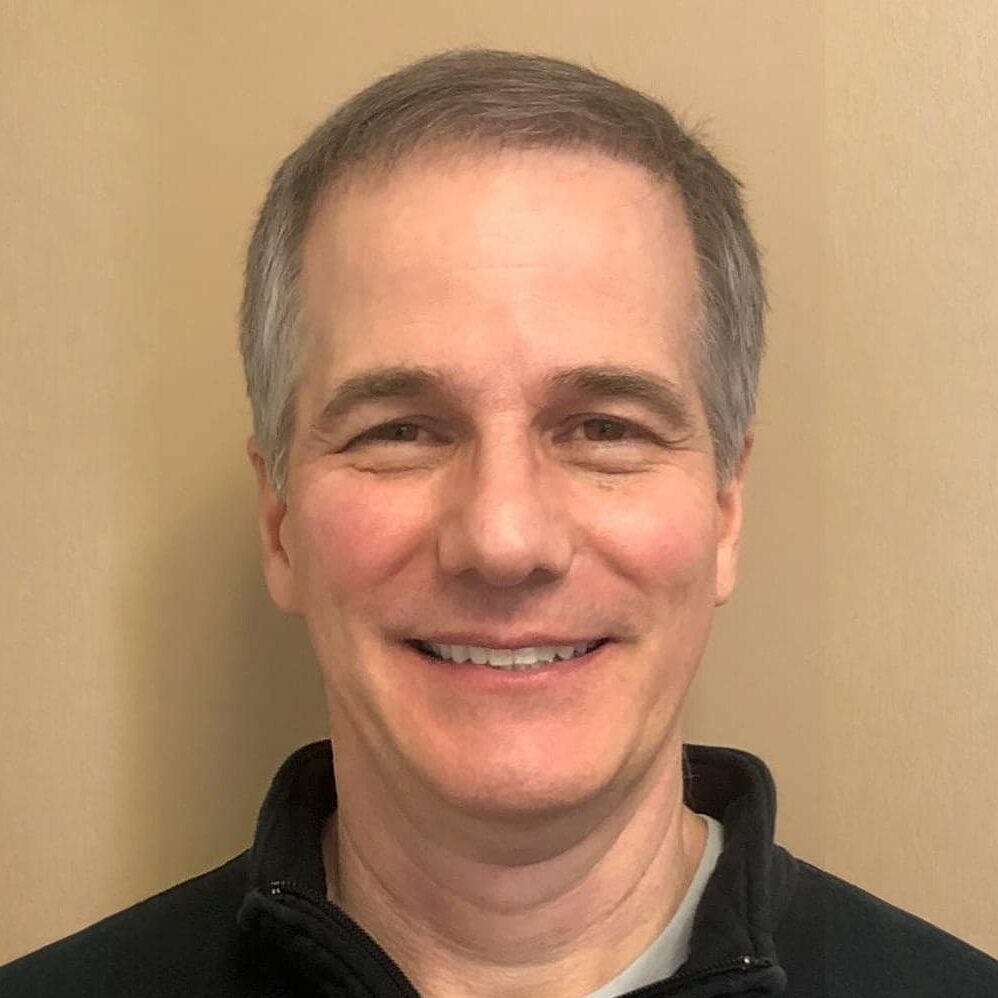 Man named Joe Nocito with short, gray hair and a friendly expression stands against a beige wall, wearing a black jacket.