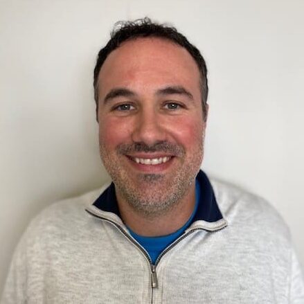 Man named Justin Malarkey with curly dark hair and light stubble smiles at the camera, wearing a light gray sweater over a blue shirt.