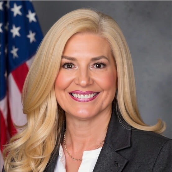 Smiling blonde woman named Paula Minichello with long hair in a dark blazer and white shirt, with an American flag in the background.