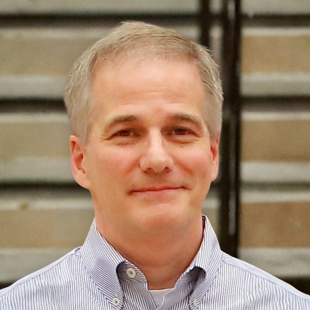 Professional headshot of Joe Nocito in a striped button-down shirt, standing in front of an industrial-style background for Minichi Inc.